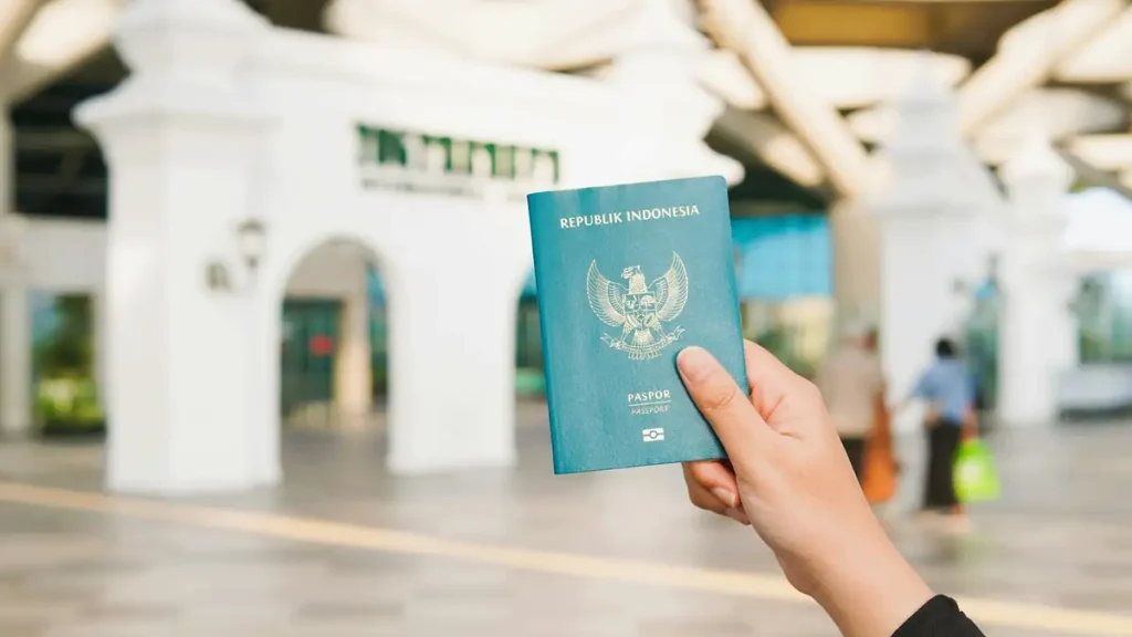 A hand holding a green Indonesian passport in front of an airport terminal